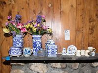 Photo showing the entire collection of blue and white ceramic items on a wooden shelf, including flower vases with artificial flowers, ginger jars, mini beer steins, small decorative plates, and assorted small ceramic mugs or containers.