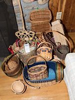 Photo showing a collection of over 20 assorted baskets in different shapes, sizes, and styles including wicker and wooden ones, some with handles, some shallow and flat, some deeper. The baskets are placed against a stone wall and wood panel background.
