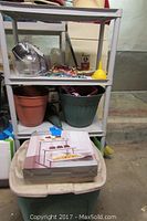 White plastic four-shelf storage unit holding gardening items including pots and watering can, with a box containing a three-tier serving stand placed in front.