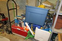 Photo showing red Coleman cooler, black metal hand truck, blue storage bin, boxed travel cooler and other miscellaneous garage items.