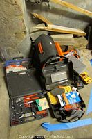 Overview photo showing Black and Decker cordless drill, drill bit case, socket sets, mouse sander, and Weller soldering gun kit on floor in garage.