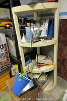 Plastic four-shelf corner unit with various small items on shelves including a blue bucket and metal scoop