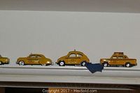 Miniature yellow taxi models lined up on a shelf showing vintage and classic styles including a pick-up style taxi and VW Beetle.