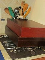Two wooden boxes stacked containing flatware sets, with some stainless steel pieces visible on counter beneath.
