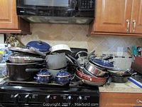 Wide view of assorted pots and pans including enamel coated and stainless steel, stacked on stove and counter.
