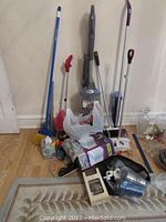 Photo showing various cleaning tools including brooms, mops, vacuum cleaner, and cleaning pads grouped on a carpeted floor against a wall.