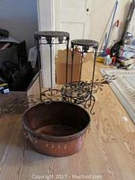 Photo showing two metal plant stands and large copper basin on wooden floor in cluttered room.