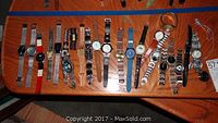 Photo showing a collection of approximately 20 assorted men's and women's wristwatches laid out on a wooden surface. Varied styles, colors, and bands visible.