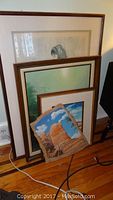 Multiple framed artworks and original art grouped leaning against wall on wooden floor