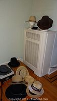 Eleven men's hats arranged on and next to a white heater cover, including straw, felt, and leather in various colors