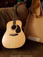 Acoustic guitar resting on a couch next to an open cardboard box showing original packaging.