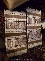 Photo of two handmade wool carpets with geometric ethnic patterns and fringed edges laid side by side on a dark patterned carpet.