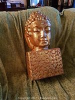 Large gold-colored Buddha head sculpture resting on green fabric. Noticeable nose damage with white coloration on the nose.