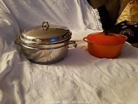 Two pots on white fabric background; stainless steel Hackman pot and orange La Creuset pot.