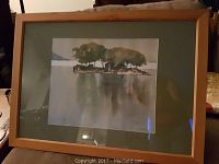 Framed Lou Charton watercolour painting depicting an island with trees and water reflections, light wooden frame and matting present.