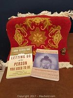Red wool prayer carpet with gold patterns, two Buddhist-themed books, and a small brass Buddha statue on a brown surface.