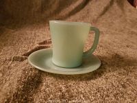 Photo of a pale green Jadeite glass cup and saucer set on a textured brown surface, showing the cup with handle and the matching saucer beneath it.