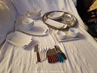 Photo showing white ceramic serving dishes, small bowls, an oval silver-colored serving tray, and assorted serving utensils with various handles.