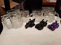 Four clear Batman design collectible glasses and three Batman-themed die-cast cars arranged on a white surface