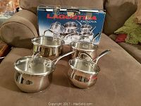 Eight-piece Lagostina stainless steel cookware set including various sized pots, saucepans, and lids displayed on a couch with original box in background.
