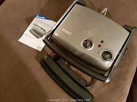 Breville Gourmet Health Grill closed, showing front stainless steel surface with dials and indicator lights, alongside the included manual.