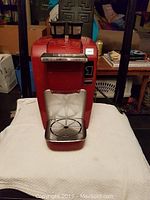 Front view of the red Keurig coffee maker showing the drip tray, control buttons, and top pod insertion area on a white textured surface.