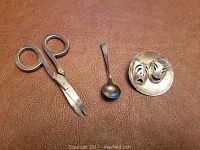 Three items on a brown surface: scissors, spoon, and brooch with theater masks