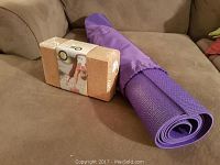Photo showing a rolled purple yoga mat with a purple strap and a cork yoga brick in packaging placed on a brown couch.