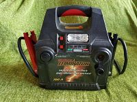 Front view of Motomaster Eliminator Mobile Booster Pack 700A battery charger showing handle, clamps, control buttons, indicator lights, and flashlight.