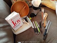 Set of kitchen items including wooden bowl with utensils, cookbook, coffee pot, knives, and kitchen tools arranged on a couch.