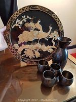 Photo of round Asian wall plaque with gold-tone peacock and floral design, positioned behind a black sake set with one bottle and four cups on a glass surface.