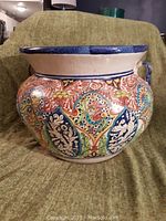 Large ceramic urn on green fabric background, detailed front view showing colorful floral/leaf motif and blue rim
