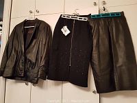 Three clothing items hanging on closet doors: black leather jacket, black skirt with studs and zipper, and a black leather-like skirt with tags.