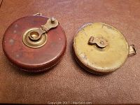 Photo showing the front side of the two round measuring tapes, one in leather case with brass winding component, the other in full brass case with locking clip