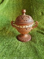 7-inch Eurasian decorative candy dish on green fabric background, showing front view with lid and symmetrical handles, intricate carved floral and geometric patterns all around.