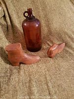 Three items on fabric background: a brown glass jug embossed with 'Javex' and two wooden whittled objects resembling a boot and a shoe.