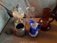 Full view of the lot showing oil lamp, hurricane shade, creamer, amber pitcher, metal canister, and floral frog on a sofa.