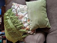 Photo showing a green woven throw blanket with fringes, stacked with two decorative pillows on a couch.