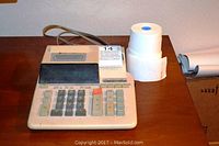 Front view of beige Sharp electronic printing calculator with keypad and two paper rolls on a wooden table.