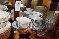 Stacked fine china with dinner plates, salad plates, saucers, bowls, and tea cups on a wooden surface. Glassware items visible beside them including clear serving bowls, salt shaker, candle holder, napkin holder, butter dish, spoon rest.