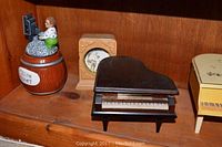 Wooden shelf displaying a brown music box with a girl figurine playing piano on top of drum base, a small wooden clock music box, and a black miniature grand piano figurine.