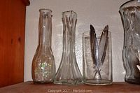 Three glass vases of varying height and shape on a wooden shelf against a white wall.