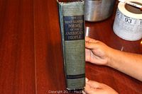 Side spine view of the green hardcover book titled 'The Best Loved Poems of the American People' held upright by a hand on a wood table with paint cans in the background.
