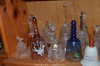 Shelf holding 9 assorted glass and crystal bells in a variety of styles, colors, and sizes. Includes amber, clear, blue, and white decorative bells with different shapes and handles.