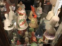 Full display shelf with multiple collectible decorative bells of different styles and sizes, showing hand-painted designs, animal figurines and holiday themes.