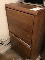 Wood finish laminate file cabinet with two drawers against a wall, showing some surface wear.