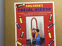 Photo of the cardboard box packaging showing the label for the children's cheval mirror with colorful pencil illustrations and a picture of a child looking into the mirror.