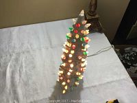 Front view of the white ceramic Christmas tree lit up with multi-color bulbs on a white table.