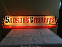 Illuminated vinyl sign displaying the text 'Season's Greetings' glowing brightly on a dark background.
