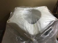 White plastic elevated toilet seat still in original plastic wrapping, showing the seat's opening and thick construction.